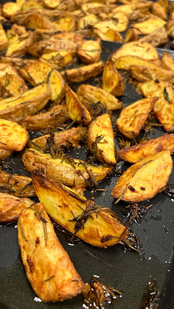 Golden roasted skin-on potato wedges with herbs on a dark baking surface.